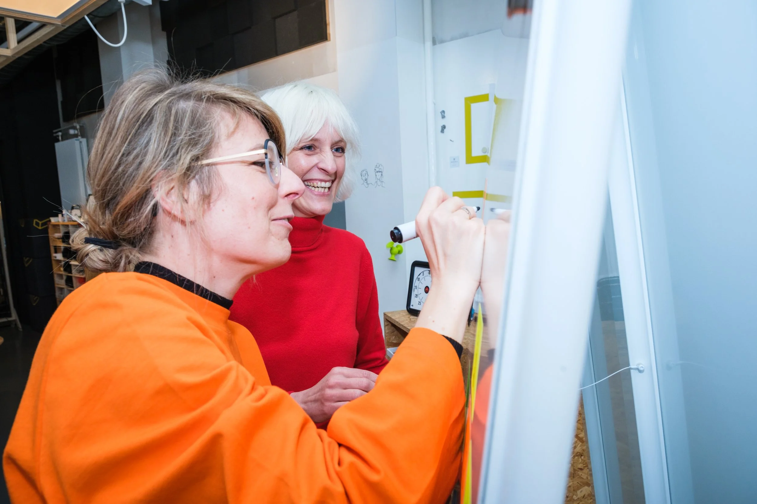 Zwei Frauen, eine schreibt auf einer weißen Tafel, die andere lächelt, im Hintergund eine Uhr und Notizen an der Wand
