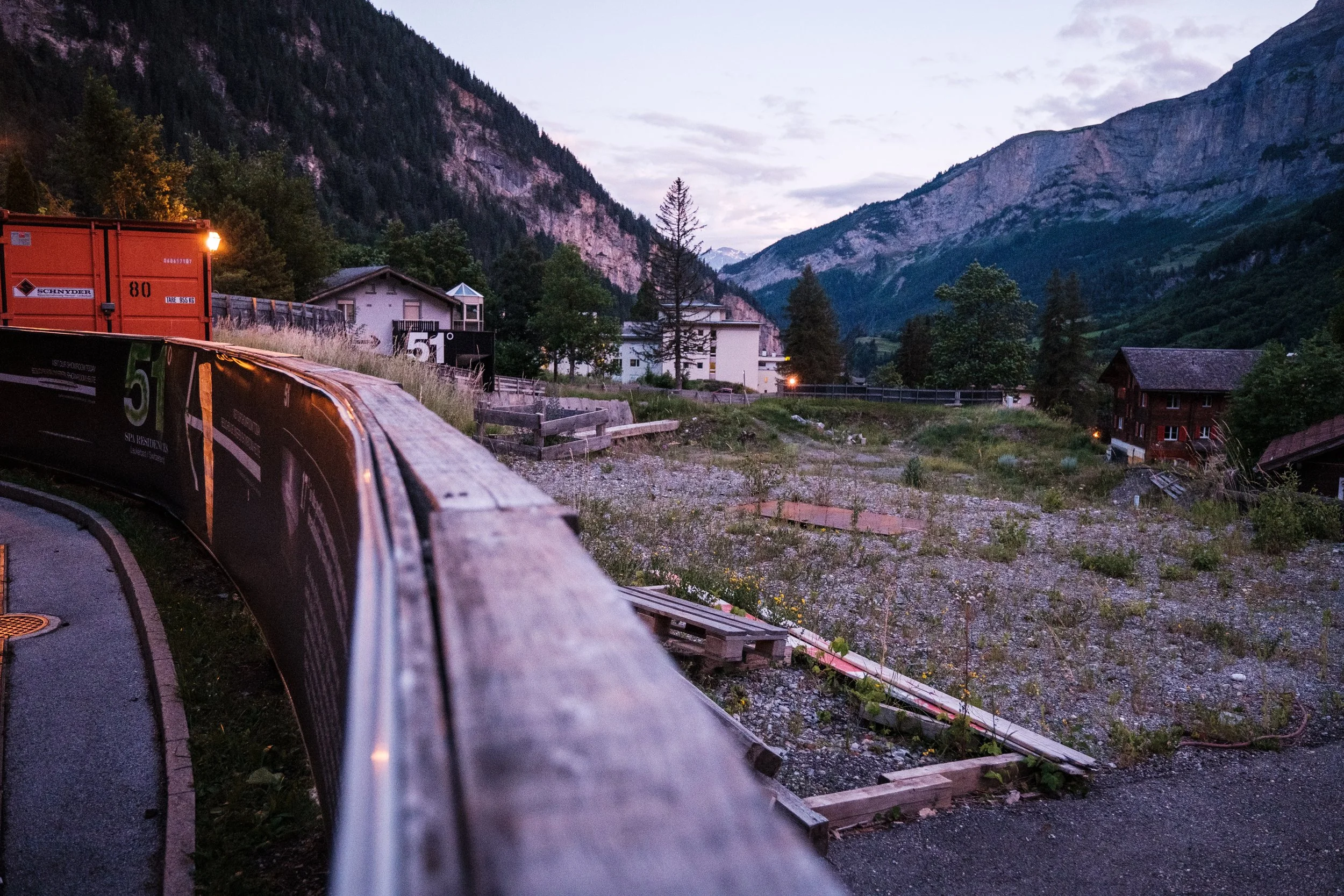Blick von einem Bahnsteig auf eine Bahnstrecke, Häuser, Bäume und Berge bei Sonnenuntergang oder Dämmerung in einer ländlichen Gegend in den Alpen.