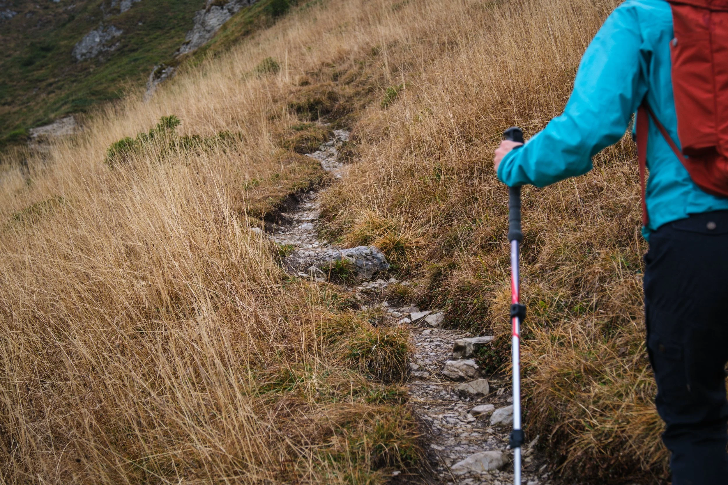 Person mit blauer Jacke und rotem Rucksack beim Wandern an einem steilen Hügel mit trockenem Gras und einem schmalen Weg.