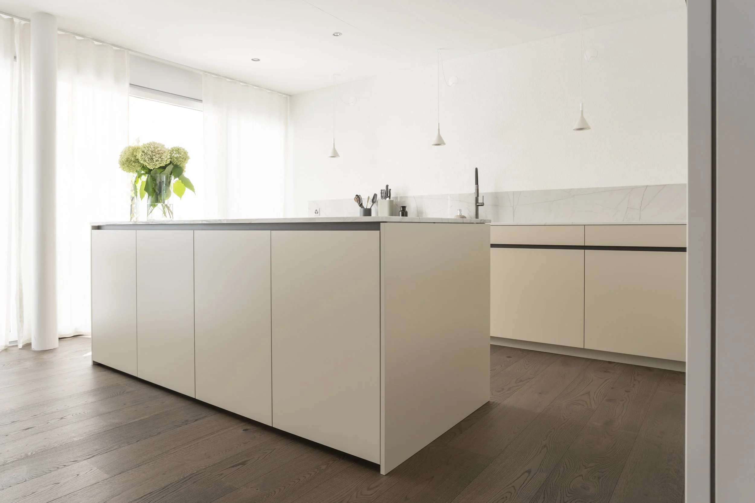 Moderne, minimalistische helle Küche mit weißen Schränken, Holzfußboden, großer Fensterfront mit weißen Vorhängen, Vasen mit Blüten auf der Kücheninsel, Deckenleuchten, Wasserhahn und Küchenutensilien an der Wand.
