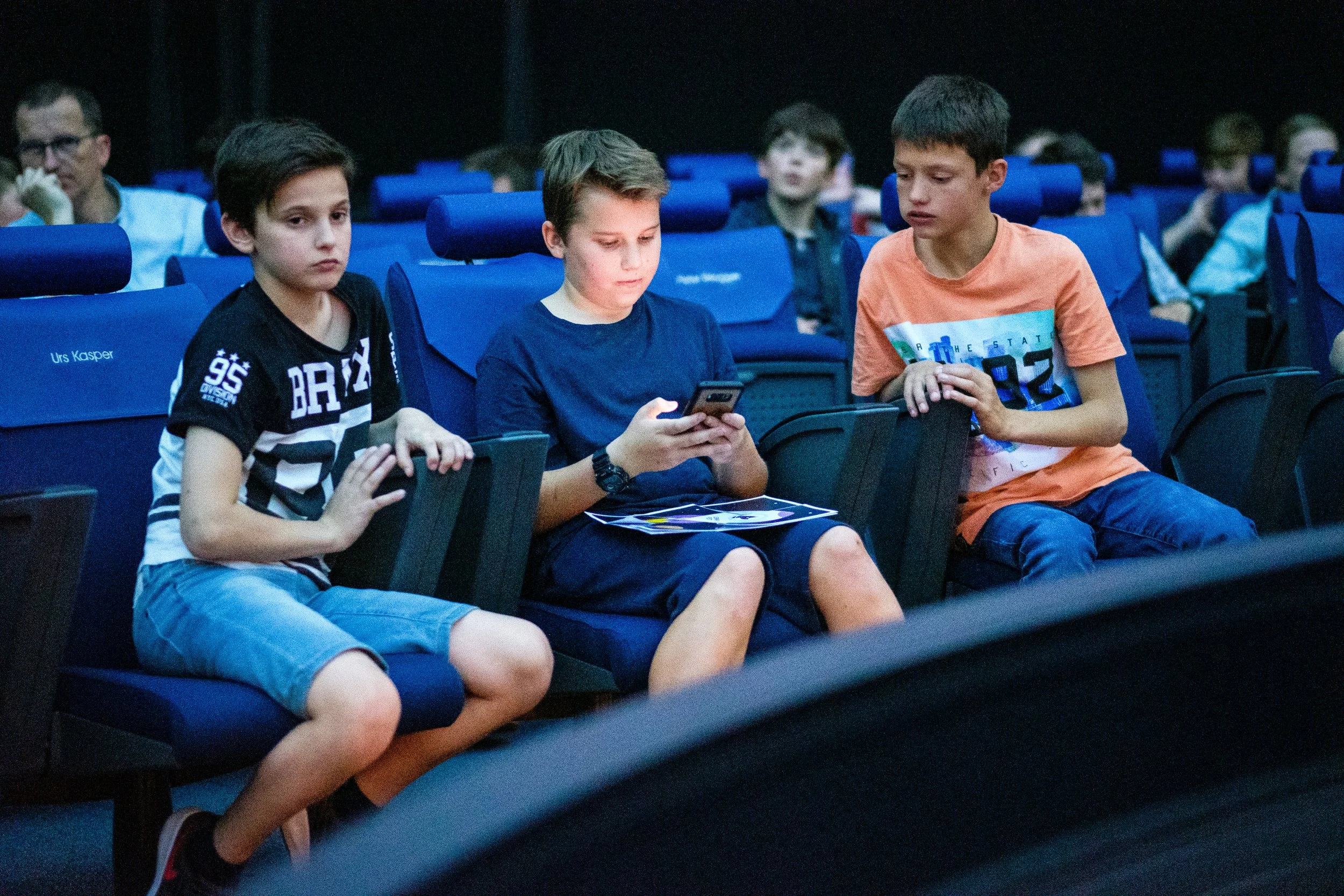 Drei Jungs sitzen in einem dunklen Theaterraum auf blauen Sitzen. Eine der Jungs schaut auf sein Handy, die anderen beiden schauen nach vorne. Im Hintergrund sind weitere Kinder zu sehen.