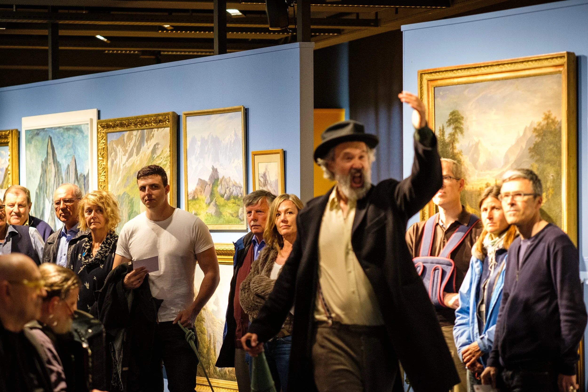 Ein Mann mit Zylinderhut und Mantel unterhält eine Gruppe von Menschen in einer Kunstgalerie mit Landschaftsbildern.