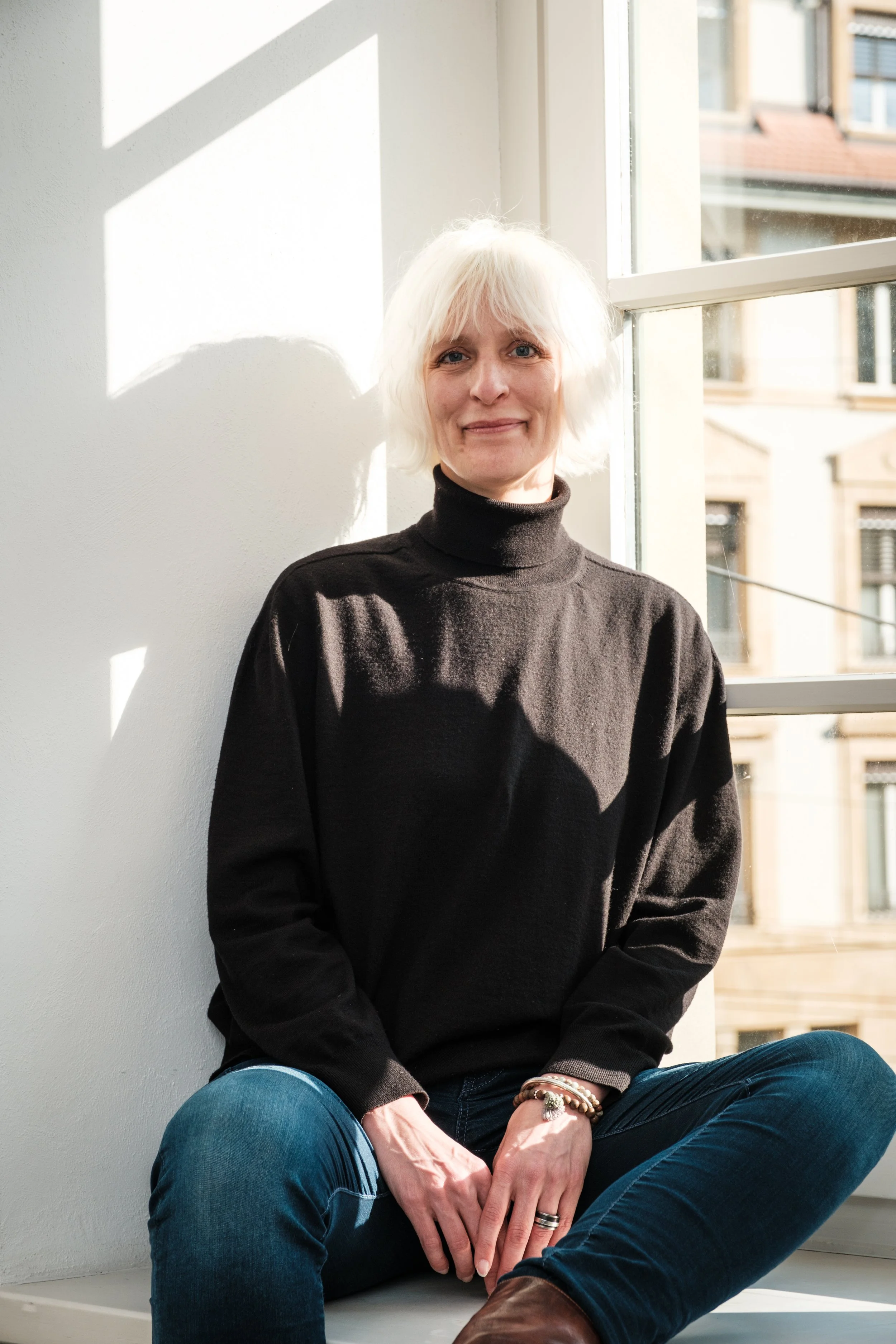 Eine ältere Frau mit kurzen weißen Haaren sitzt an einem sonnigen Tag neben einem Fenster in einem weißen Raum, trägt eine schwarze Turtleneck und blaue Jeans.
