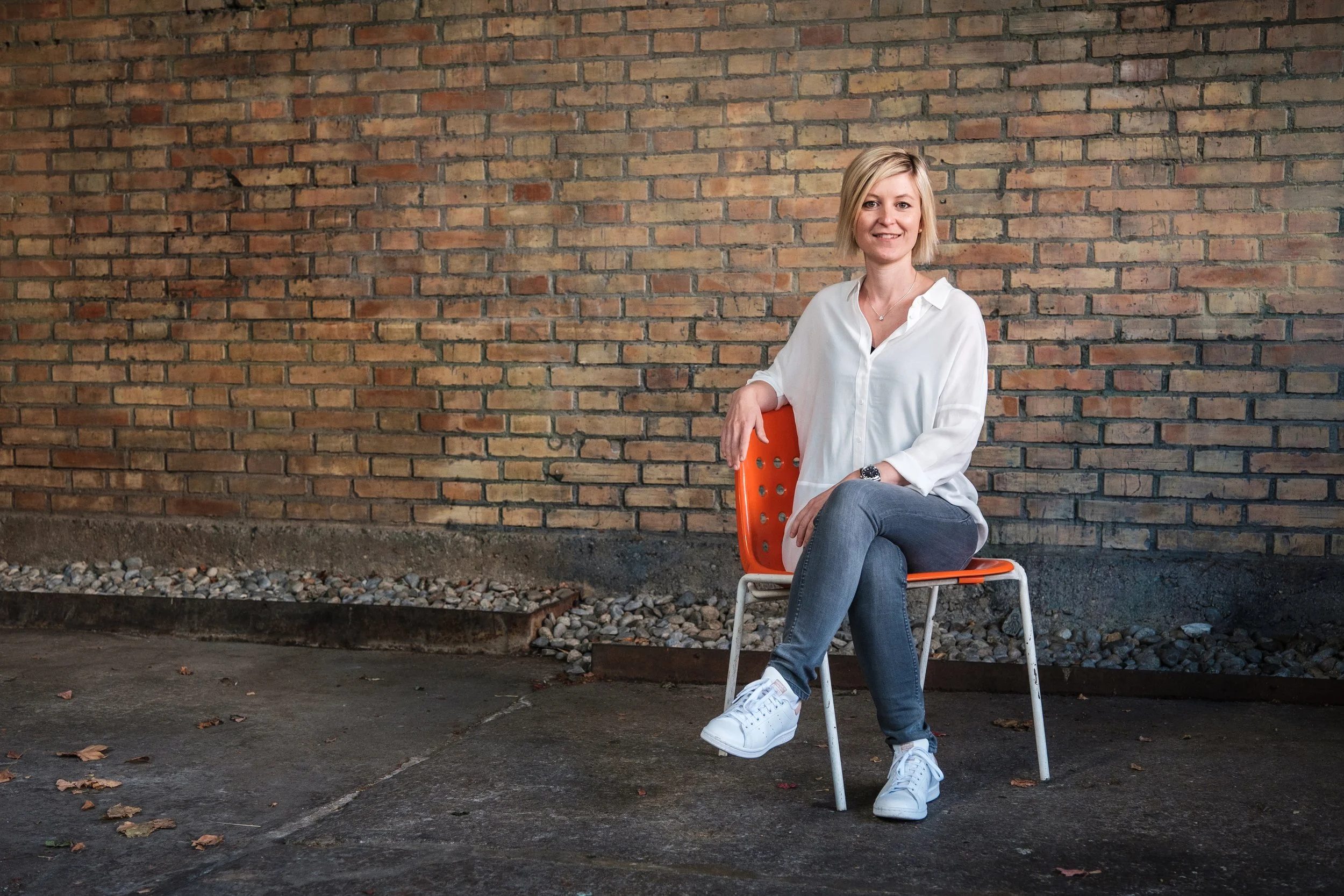 Junge Frau sitzt lächelnd auf einem orangefarbenen Stuhl vor einer Ziegelmauer, trägt eine weiße Bluse, Jeans und weiße Turnschuhe.