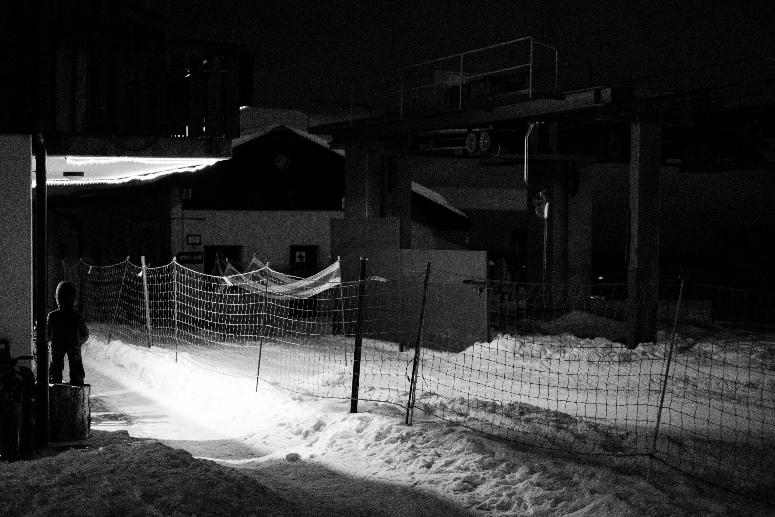 Ein Kind steht im Schnee bei Nacht, umgeben von Schneeflächen und einer Schneeschutzzaun, dunkle Gebäude im Hintergrund, schwaches Licht auf dem Schnee