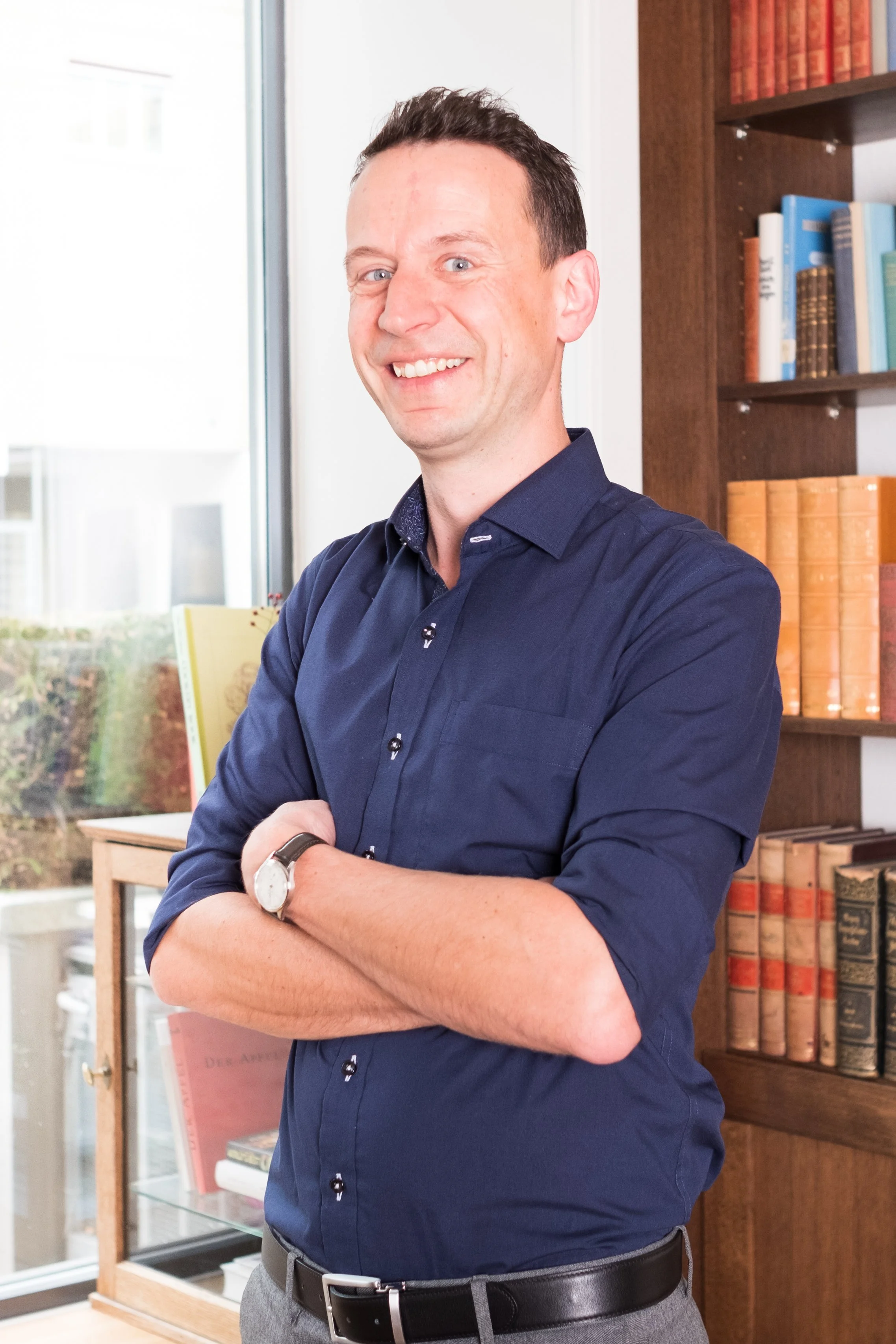 Ein Mann mit dunklem Haar, blauer Hemd, in einem Raum mit Bücherregalen und Fenster im Hintergrund.
