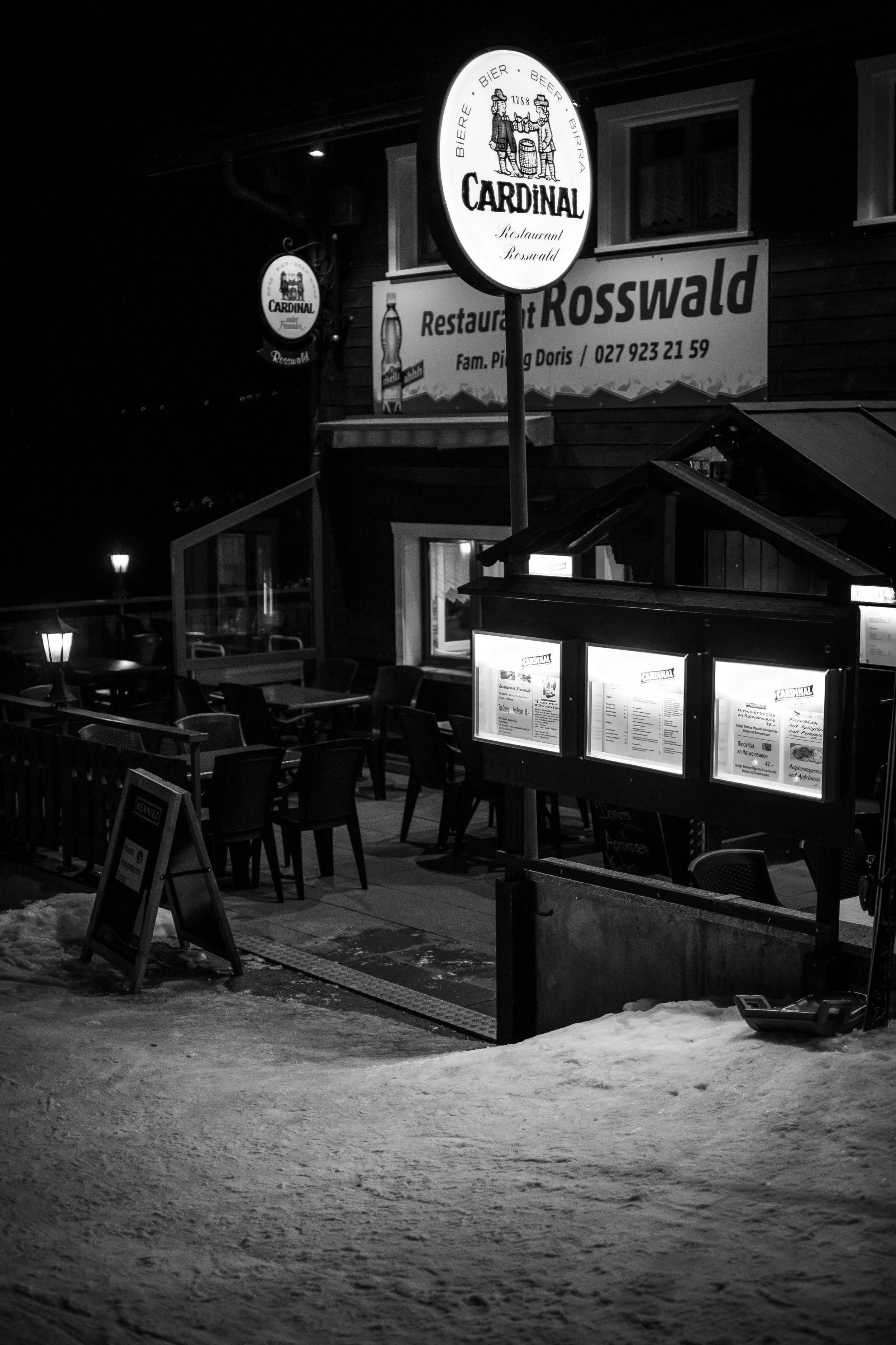 Dunkle, leere Terrasse eines Restaurants namens Rosswald bei Nacht, mit beleuchteten Anküdigungstafeln, einigen leeren Tischen und Stühlen, Schneematsch auf dem Boden und einem großen, runden, beleuchteten Schild mit dem Logo von Cardinal Bier.