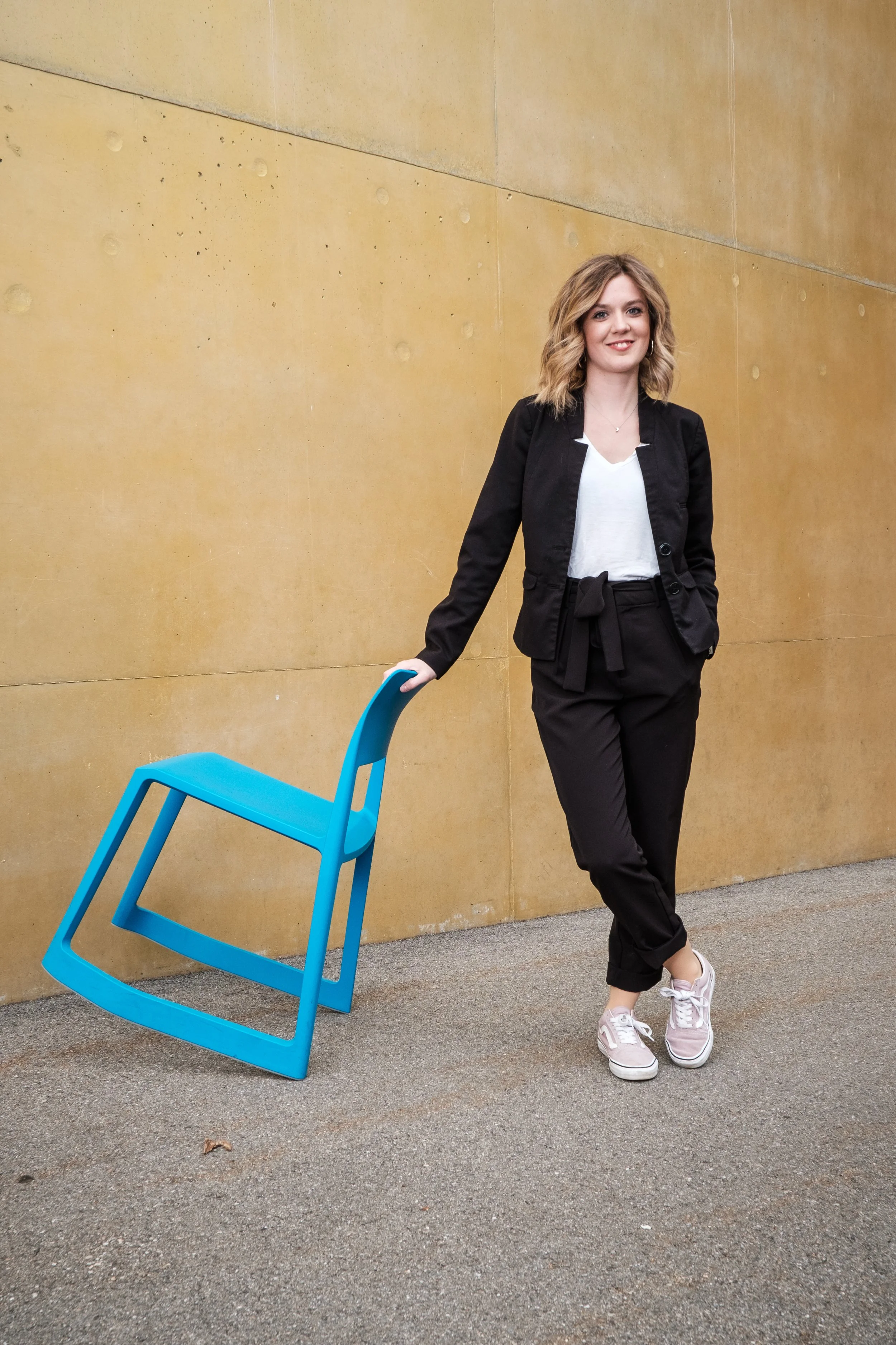 Junge Frau steht an einer Wand mit blauer Stuhl, lächelt, trägt schwarze Hose, weiße Bluse und schwarze Jacke, trägt Turnschuhe.