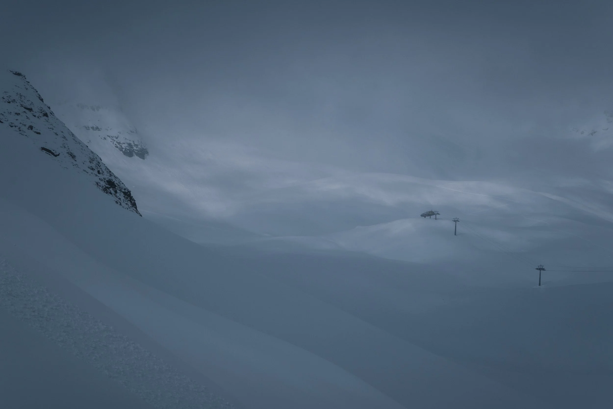 Schneebedeckte Berge und eine Skipiste mit Skilift in einer winterlichen Berglandschaft, grau-bedeckter Himmel.