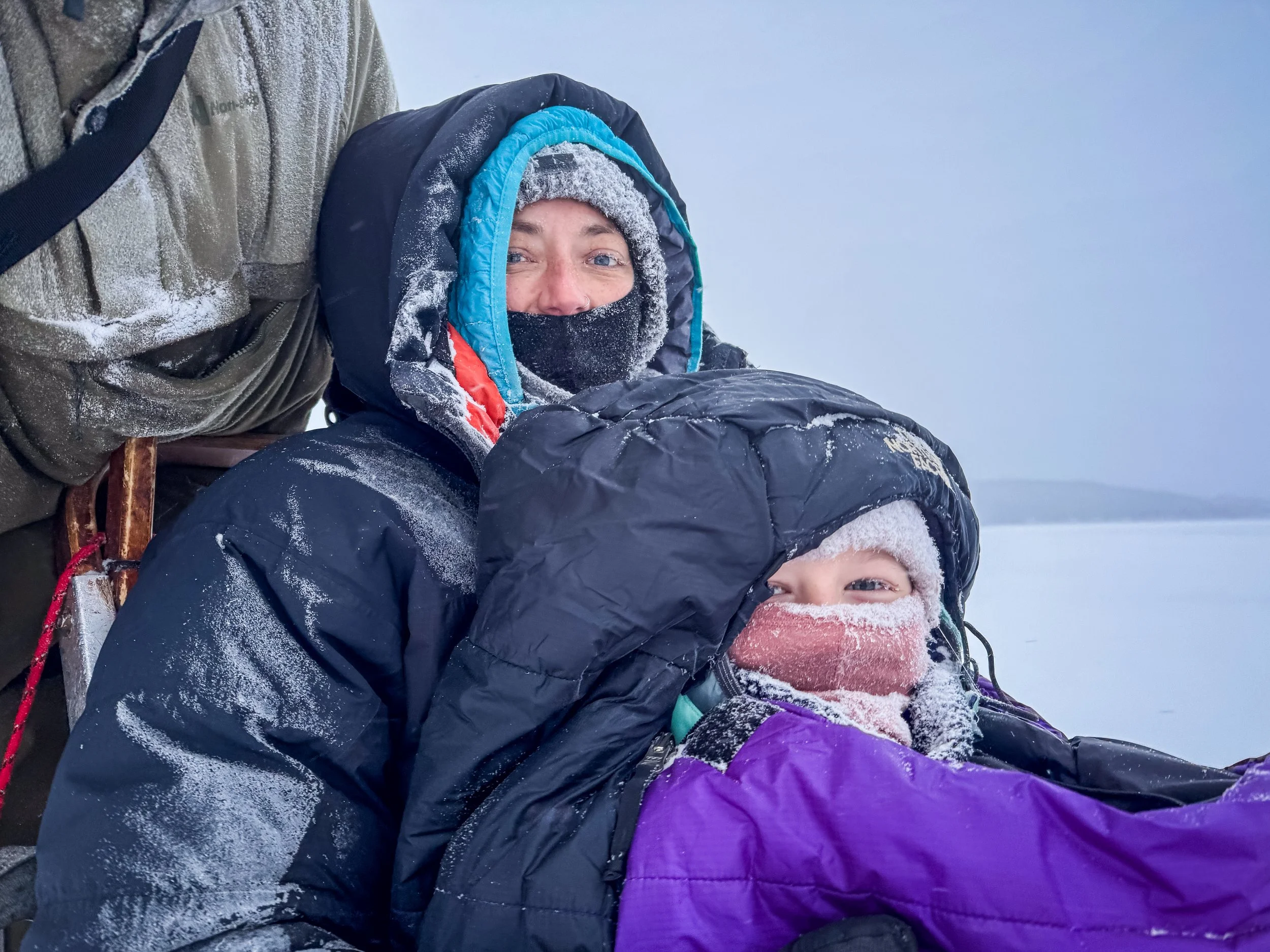 Zwei Personen in Winters Kleidung im Schnee, eine Person sitzt und die andere steht hinter ihr, beide tragen Mützen und Gesichtsmasken, um sich vor der Kälte zu schützen.