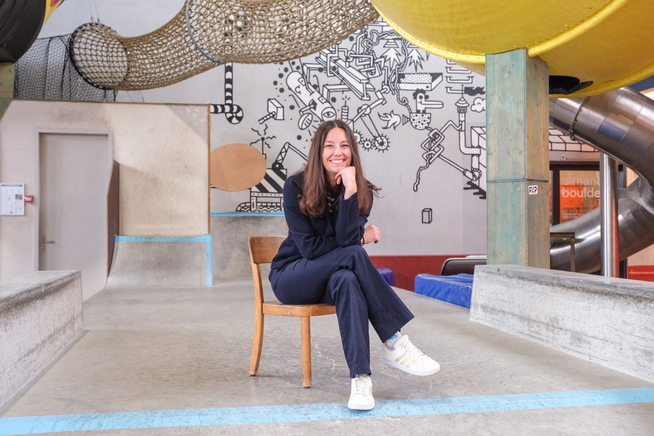 Junge Frau sitzt auf einem Stuhl in einem Indoor-Spielplatz mit Graffiti-Wand im Hintergrund, trug schwarze Kleidung und weiße Turnschuhe, lächelnd.