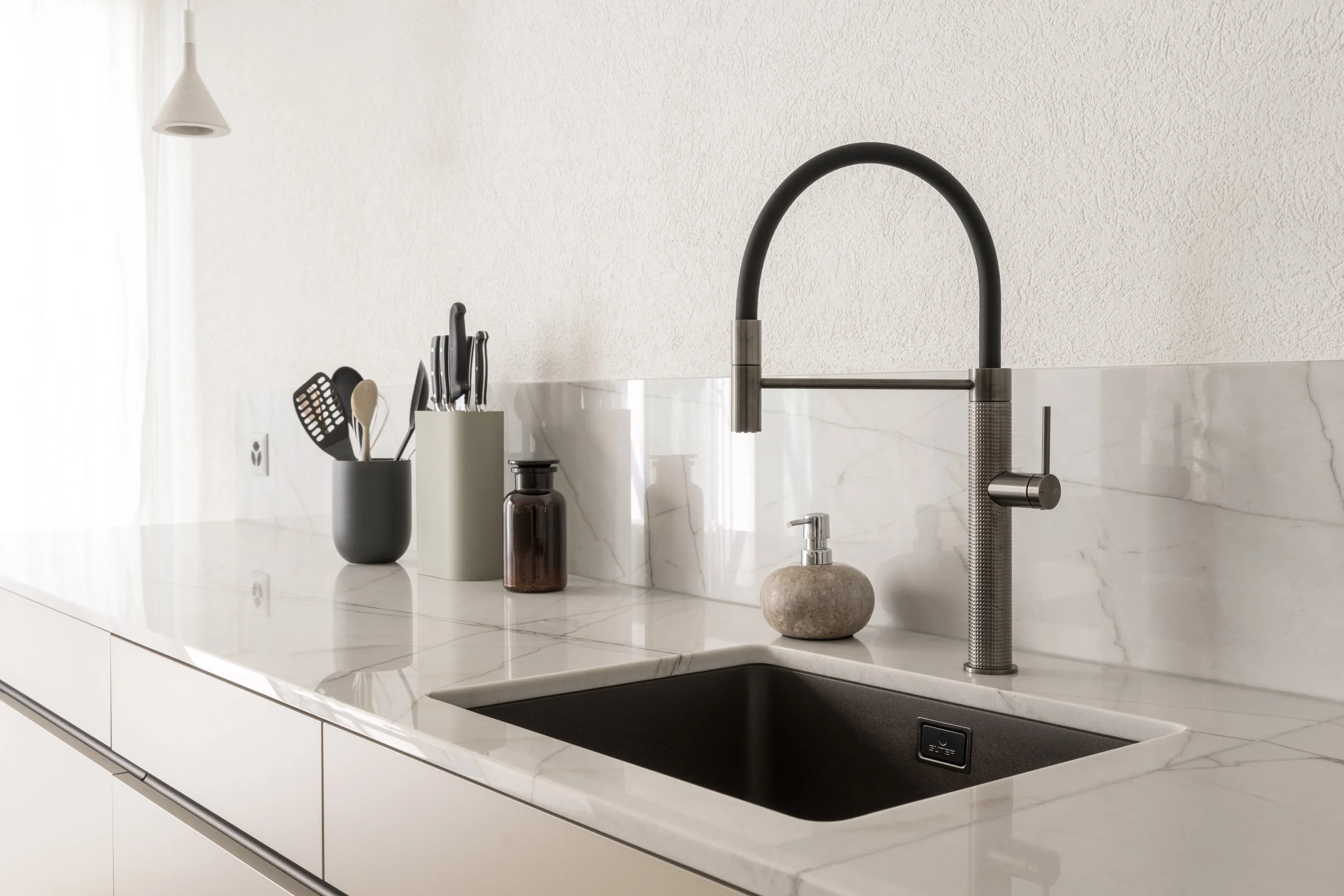 Moderne Küche mit weißem Marmor-Arbeitsplatten und schwarzer Spüle, Wasserhahn aus Edelstahl, Küchenutensilien in schwarzen und grauen Behältern, Seifenspender aus Stein, helle Wände mit Textur.