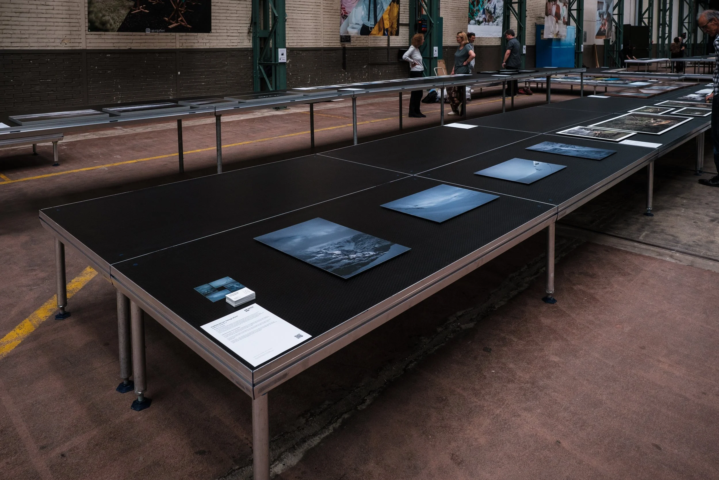 Ausstellungstisch mit Fotografien und Informationsmaterial in einer Galeriehalle, im Hintergrund stehen Besucher und weitere Tische.