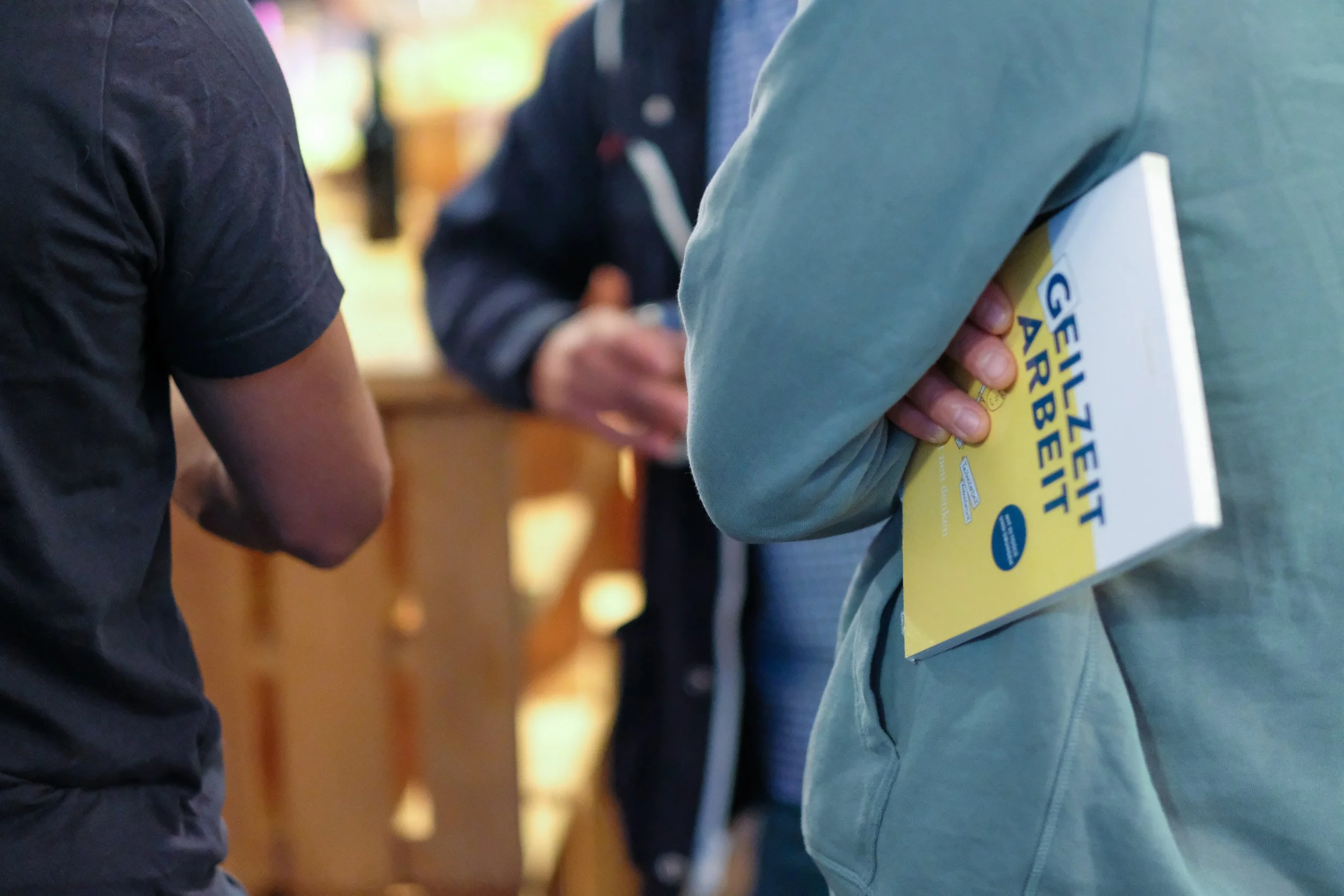 Menschen sprechen bei einer Veranstaltung, eine Person hält ein Buch mit dem Titel 'Gehe Zeig Arbeitswelt' in der Hand.