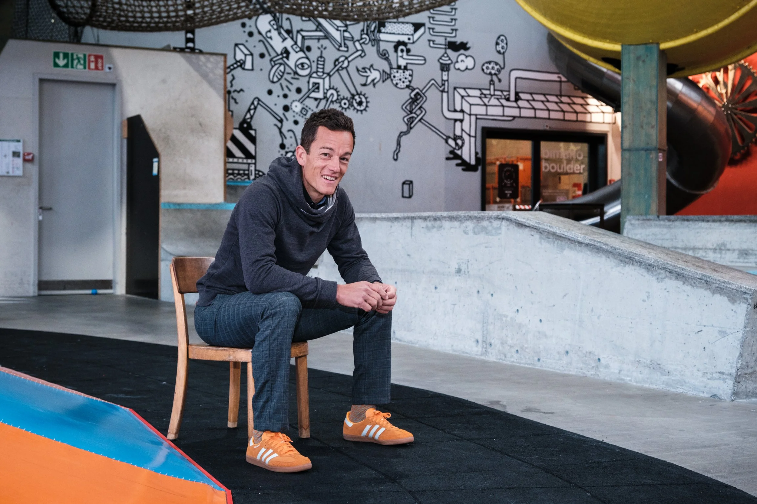 Ein Mann sitzt auf einem Holzstuhl in einer Indoor-Kletterhalle mit Graffiti-Wand im Hintergrund, trägt eine dunkle Kapuzenjacke, karierte Anzughose und orange Turnschuhe.