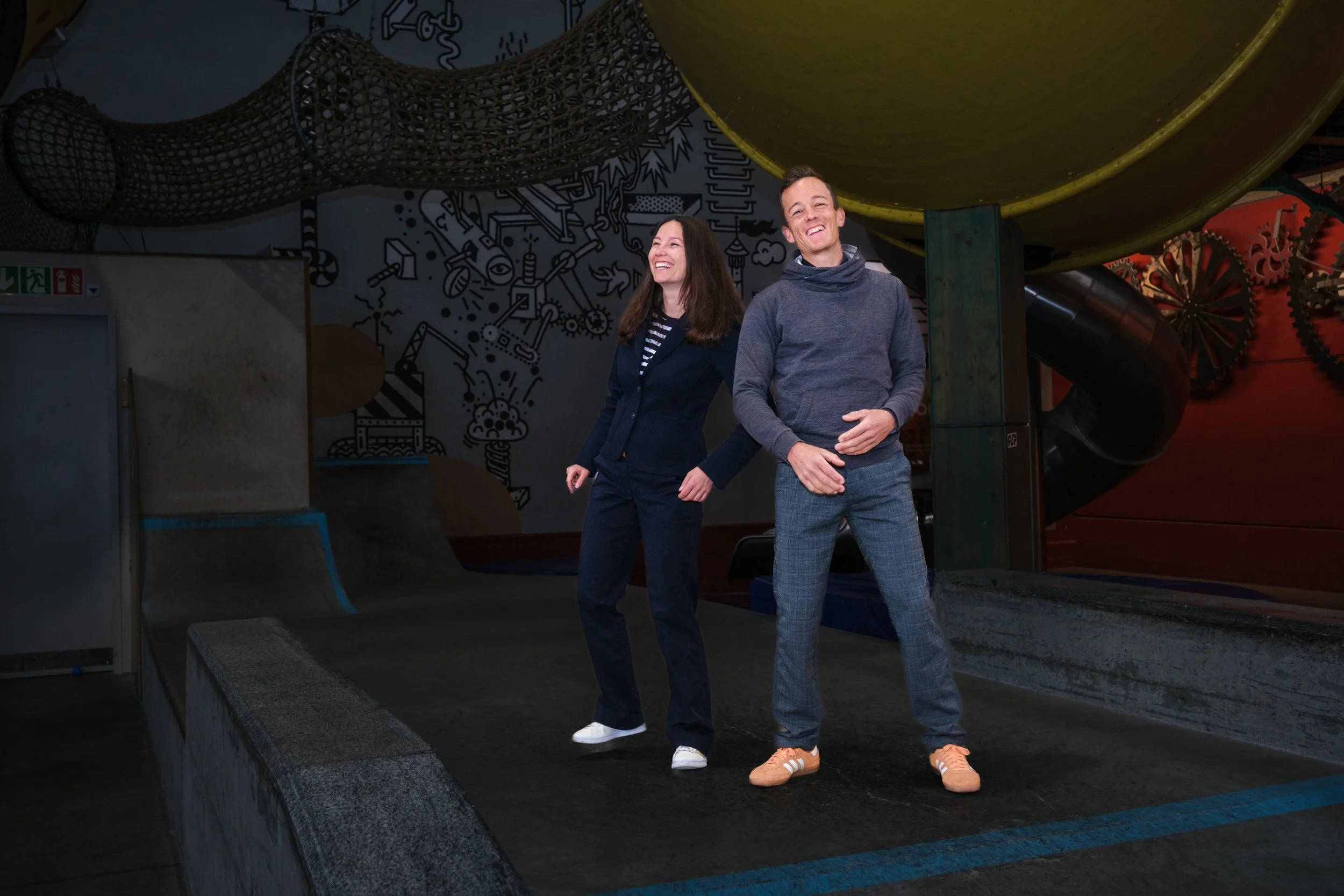 Ein Mann und eine Frau lachen und tanzen in einem Indoorspielpark, im Hintergrund Rutschen und eine Wand mit Graffiti.