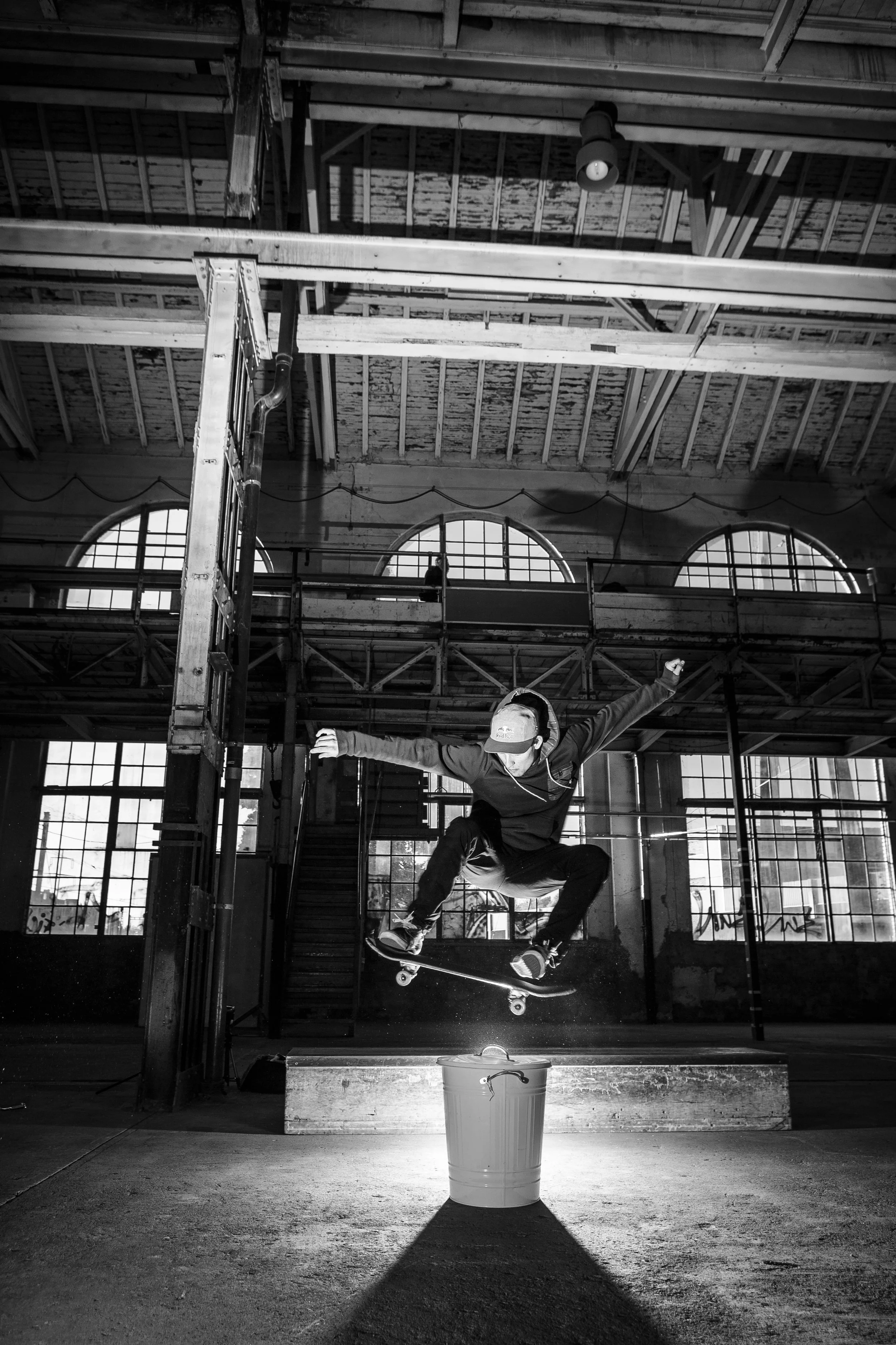 Skateboarder springt über einen Eimer in einem hell erleuchteten Industriehalle mit hohen Fenstern in schwarz-weiß