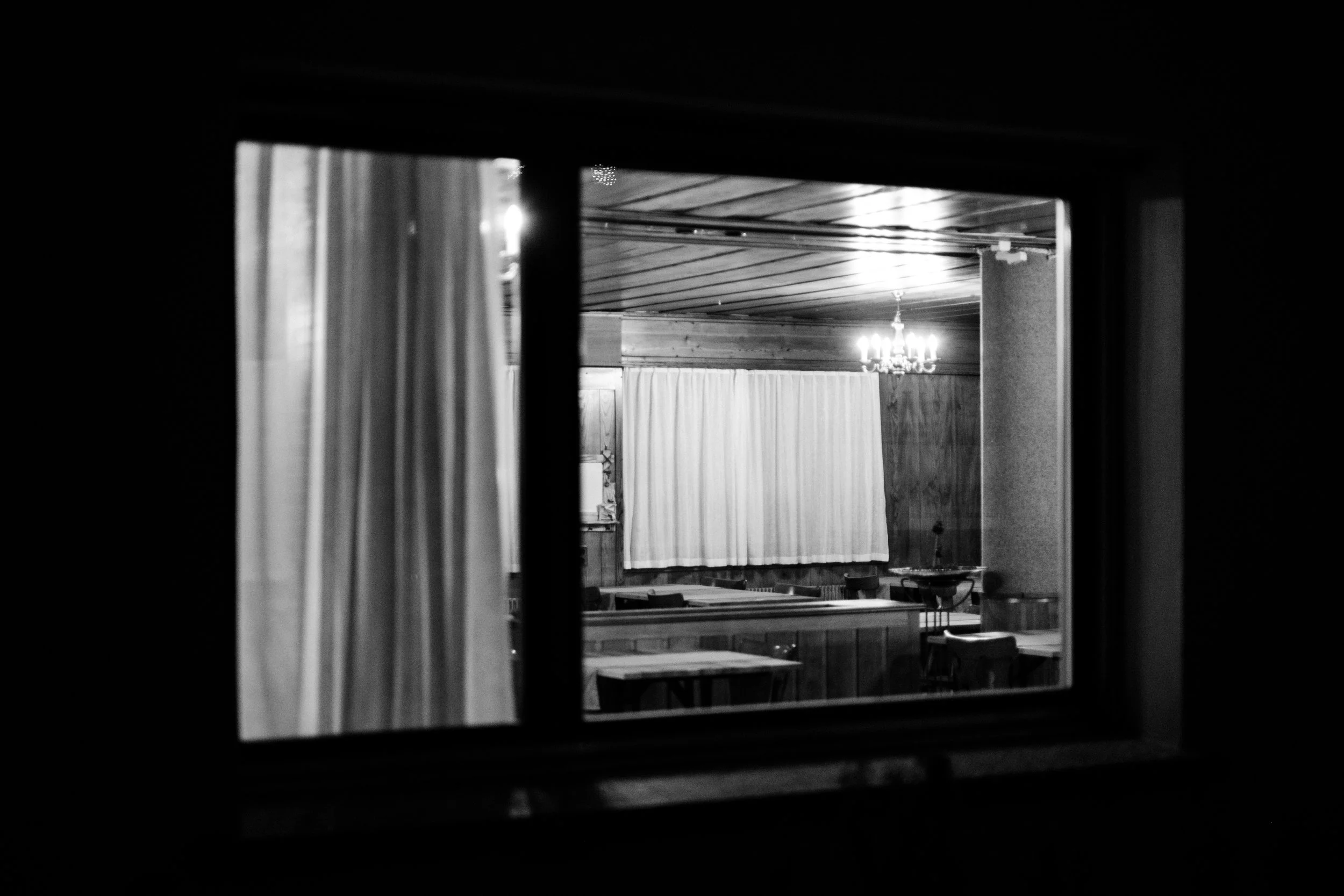 Blick durch ein Fenster auf eine kleine, leere, dunkle Restaurant- oder Salonraum mit Tischen und Stühlen, Holzvertäfelung, einer weißen Gardine und einem kleinen Kronleuchter.