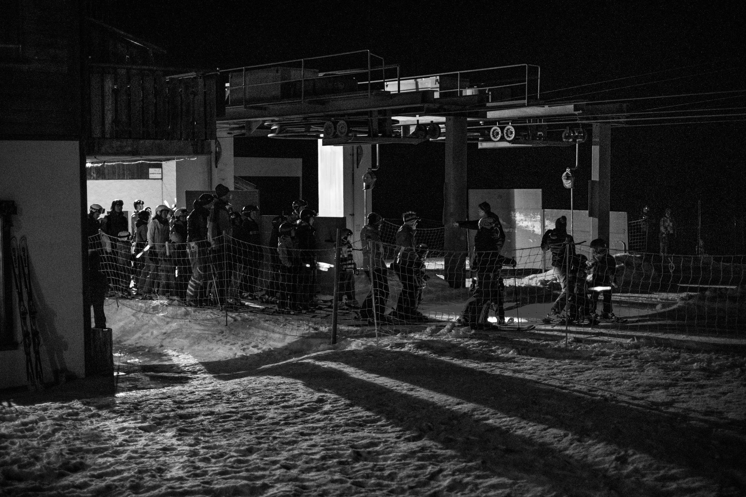 Schwarze und weiße Skifahrer warten bei Nacht an der Skilift-Auffahrt. Es ist dunkel, Schnee bedeckt den Boden, und die Skilift-Struktur ist sichtbar.