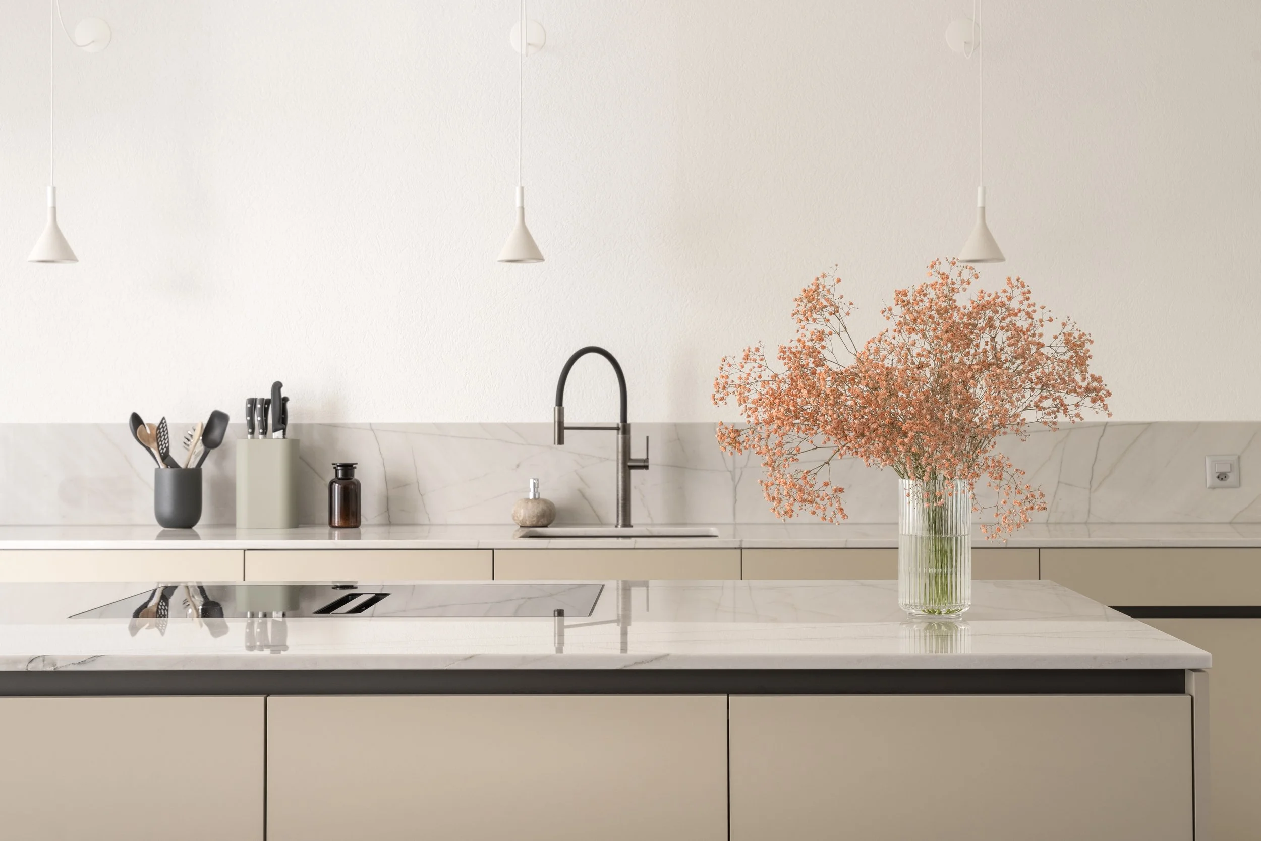 Moderner Küchenbereich mit weißen Schränken, Marmor-Arbeitsfläche, schwarzen Wasserhahn, Vase mit rosa Blumen, Kücheutensilien in Behältern und Hängelampen