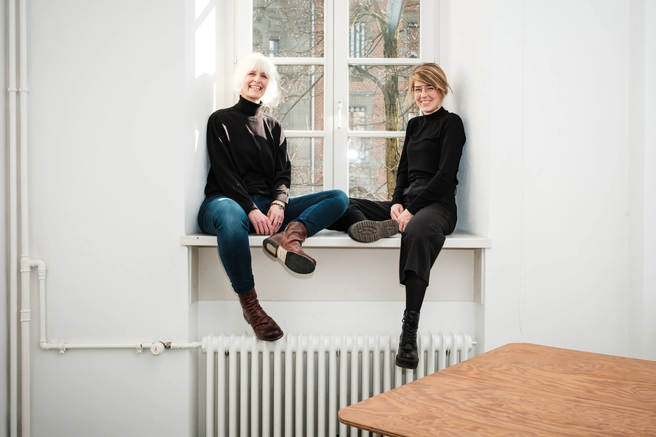 Zwei Frauen sitzen lachend auf einem Fensterbank in einem weißen Raum, hinter ihnen ein Fenster mit Blick auf Bäume und Gebäude.