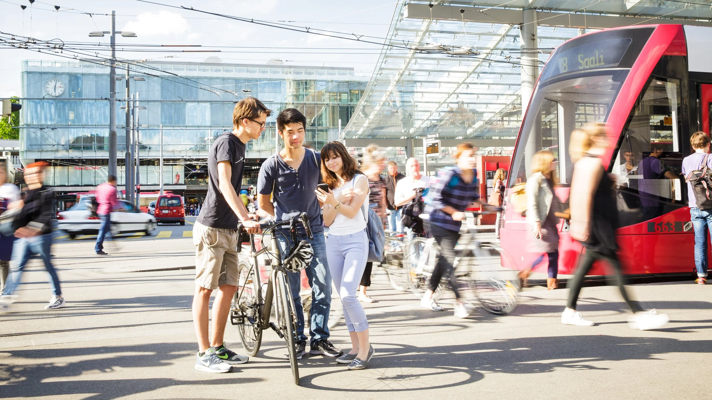 Menschen warten an einer Straßenbahnhaltestelle in einer geschäftigen Stadt, viele bewegen sich, einige schauen auf Mobiltelefone, im Hintergrund ein modernes Gebäude mit einer großen Uhr.