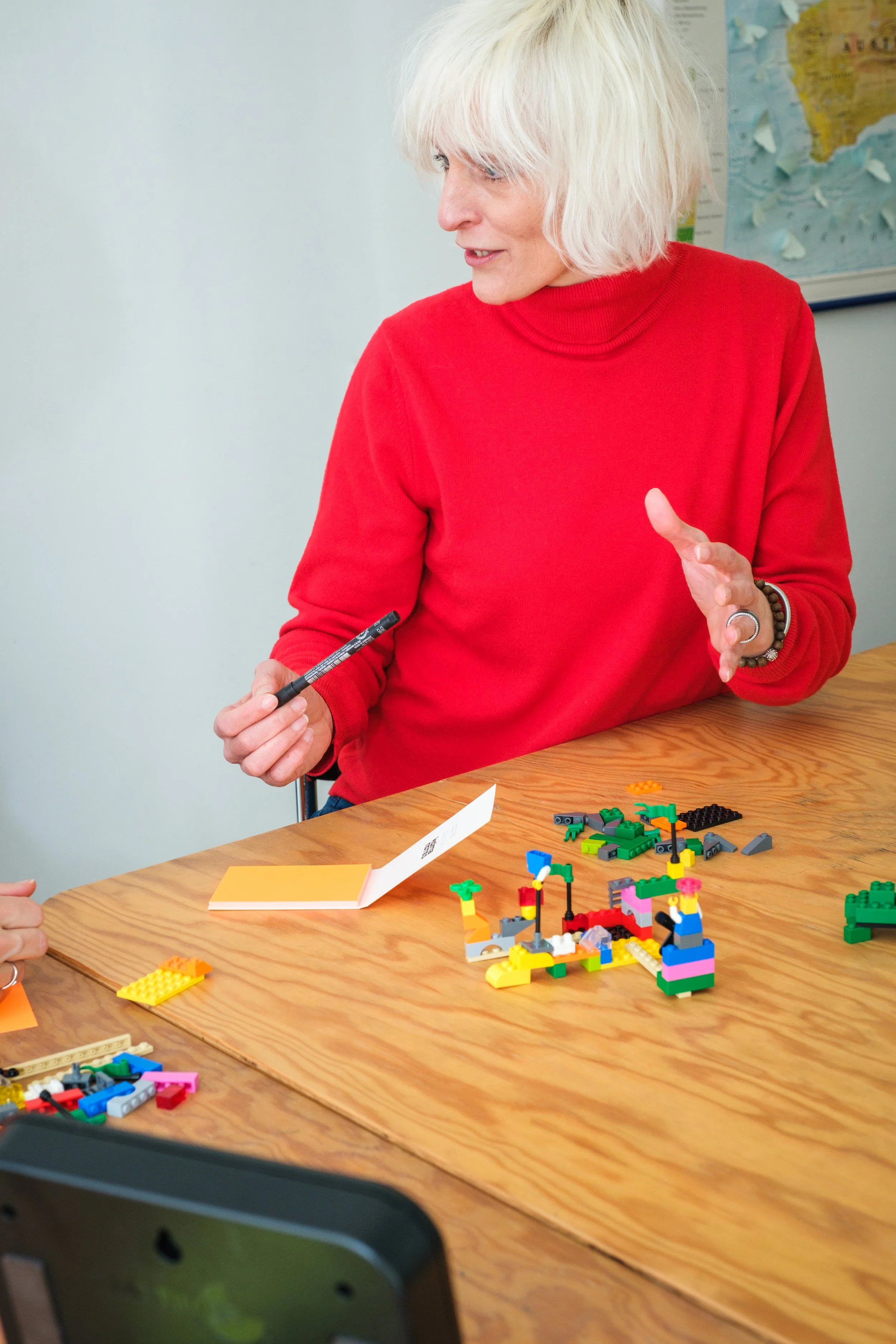 Eine ältere Frau in rotem Pullover erklärt während eines Spiels mit bunten LEGO-Steinen, die auf einem Holztisch liegen.